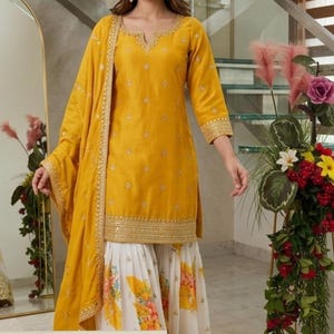 May include: A yellow kurta with gold embroidery around the neckline and hem, paired with a matching yellow dupatta featuring gold trim and white palazzo pants with a yellow and orange floral print. The text Shyam Kripaa is visible.