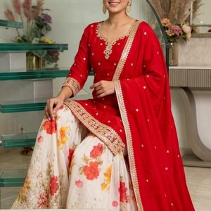 May include: A red tunic with gold embroidery around the neckline and hem, paired with a white skirt featuring a red and orange floral print. A matching red dupatta with gold trim is draped over the shoulder. The text Shyam Kripaa is visible.
