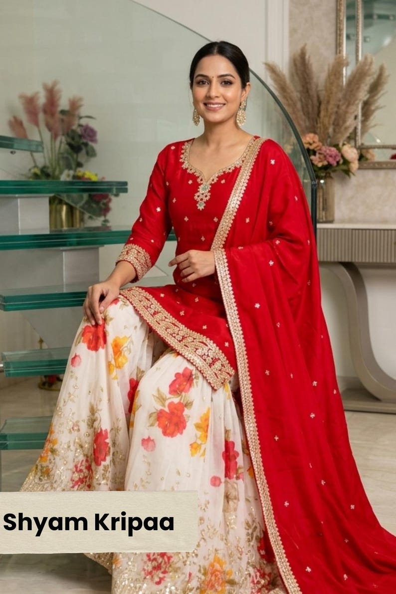 May include: A red tunic with gold embroidery around the neckline and hem, paired with a white skirt featuring a red and orange floral print. A matching red dupatta with gold trim is draped over the shoulder. The text Shyam Kripaa is visible.