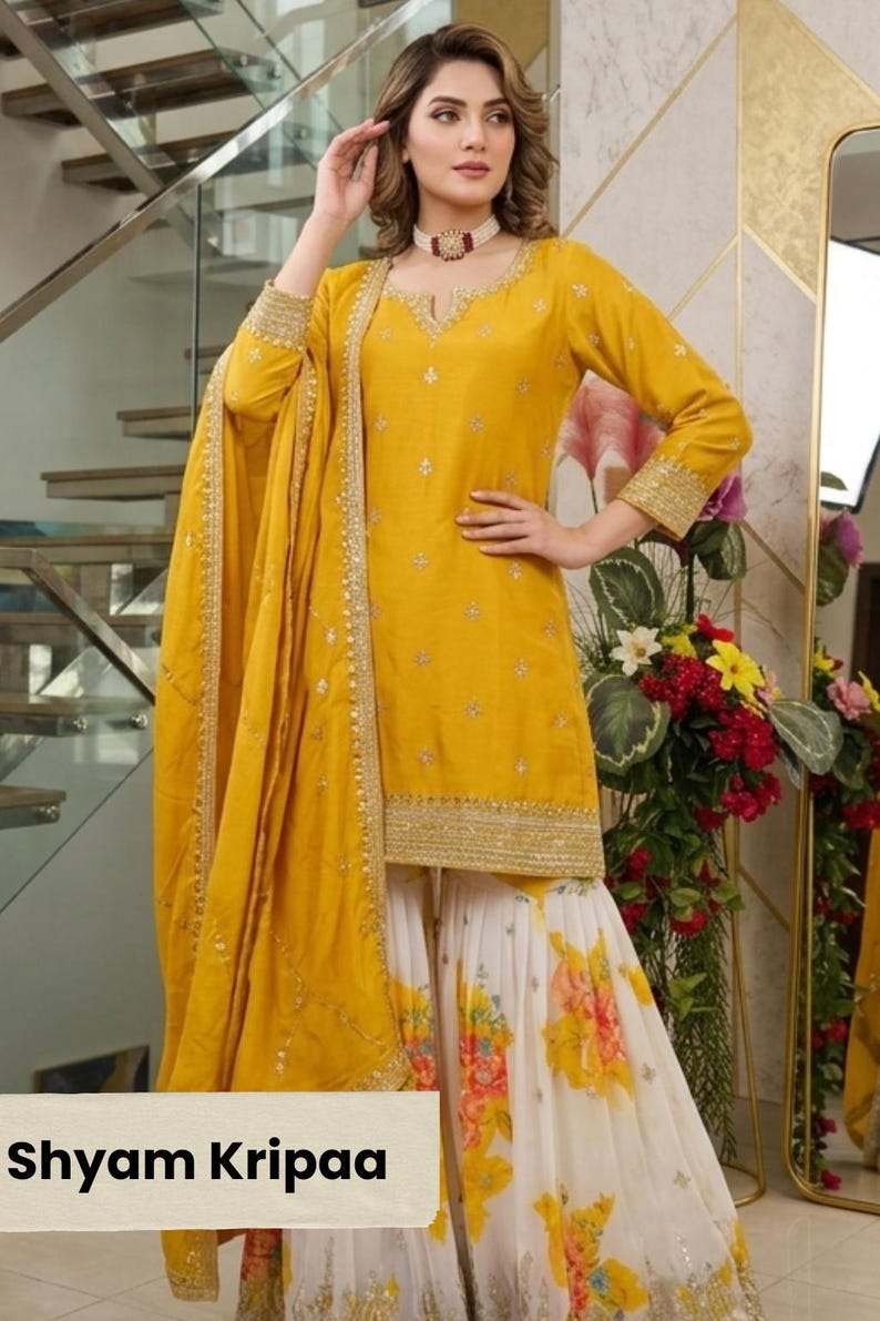 May include: A mustard yellow long-sleeved tunic with gold embroidery and a matching dupatta, paired with white wide-leg pants featuring a colorful floral print. The garment includes the text Shyam Kripaa printed on a light beige rectangular label.