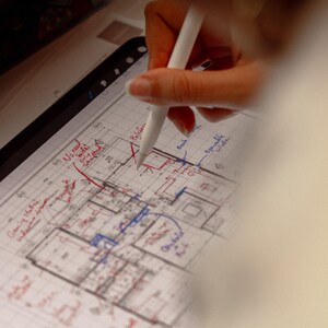 May include: A digital tablet displaying a detailed architectural floor plan with handwritten annotations in red and blue ink. A white stylus is positioned on the screen, actively marking specific areas of the technical drawing layout.