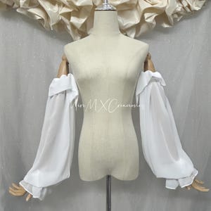May include: A pair of detachable white off-the-shoulder sleeves made of sheer, lightweight fabric. The sleeves feature a voluminous, flowing design with ruffled edges at the top and bottom cuffs. The text Miri MX Creaciones is visible in the center.