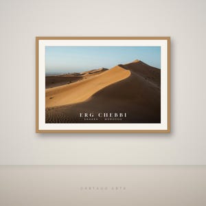 May include: A framed landscape photograph of the Erg Chebbi sand dunes in the Sahara Desert, Morocco. The image features golden-orange dunes under a clear sky, with the text "ERG CHEBBI SAHARA MOROCCO" printed in white at the bottom of the print.