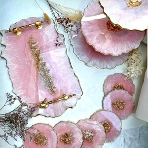 May include: A rectangular resin tray with gold handles, a tiered serving stand, and six circular coasters. Each piece features a translucent pink finish with gold leaf accents and irregular, wavy edges, creating a cohesive set of serving ware.