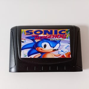 May include: A black plastic video game cartridge for the Sega 32X console. The center label features a colorful illustration of the character Sonic the Hedgehog with the text Sonic the Hedgehog 32X Edition printed in blue, red, and yellow.
