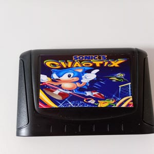 May include: A black Sega 32X video game cartridge featuring a label with the title Sonic's Chaotix in yellow and blue text. The label displays a blue hedgehog character, a green crocodile, and a small flying saucer against a dark blue background.