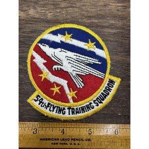 Op de afbeelding: Een ronde geborduurde patch met een witte adelaarssilhouet op een rode en blauwe achtergrond met gele sterren en een witte bliksemschicht. De tekst 54th FLYING TRAINING SQUADRON is in het zwart geborduurd. Een liniaal toont 8,9 cm breedte.