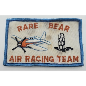 May include: A rectangular embroidered patch with a white background and a blue border. It features an orange and white airplane, a black and white checkered pylon, and the orange embroidered text RARE BEAR AIR RACING TEAM.