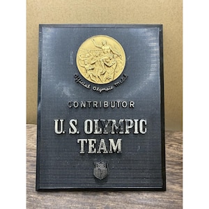 May include: A rectangular black plaque with a grid texture featuring a gold-toned circular relief of athletes. The text reads CONTRIBUTOR U.S. OLYMPIC TEAM with Official Olympic Medal below the relief and a small shield emblem at the bottom center.