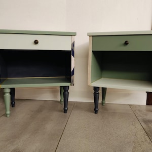 Può includere: Due comodini dotati di un cassetto con un piccolo pomello rotondo, un ripiano a giorno e quattro gambe tornite. Il comodino a sinistra presenta pannelli laterali con motivi floreali e un interno scuro, quello a destra è dipinto in verde salvia.