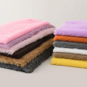 May include: A stack of rectangular faux fur fabric swatches in various colors including pink, lavender, dark brown, tan, charcoal gray, orange, white, and yellow. Each piece features a long, soft, shaggy texture with a consistent pile.