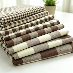 May include: A stack of folded fabric pieces featuring various brown and cream checkered patterns. The top layers display a small-scale gingham check, while the bottom layers transition into larger, bolder square patterns in alternating brown and cream.