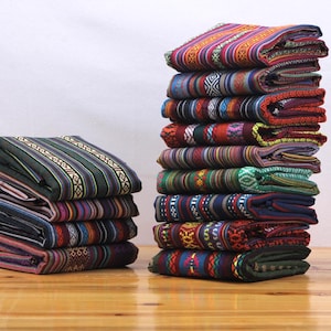Puede incluir: Una pila de diez textiles rectangulares plegados y una pila más pequeña de cuatro textiles plegados, cada uno con intrincados patrones tejidos en varios colores, incluyendo rojo, azul, verde, amarillo y morado con diseños geométricos.