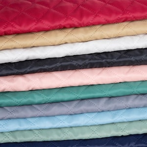 Op de afbeelding: Een stapel gevouwen stoffen met een doorgestikt ruitpatroon. De lagen zijn uitgevoerd in diverse kleuren, waaronder rood, beige, wit, zwart, lichtroze, groenblauw, grijs, lichtblauw, saliegroen en donkerblauw.