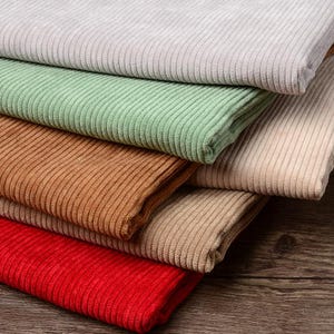 May include: A stack of folded corduroy fabric in various colors including light gray, sage green, tan, beige, and bright red. Each piece of fabric features a distinct vertical ribbed texture running across the surface of the textile material.