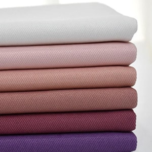 May include: A stack of six folded fabric pieces arranged in a gradient from white to light pink, dusty rose, mauve, deep berry, and dark purple. Each piece of fabric features a visible diagonal twill weave texture throughout the entire surface.