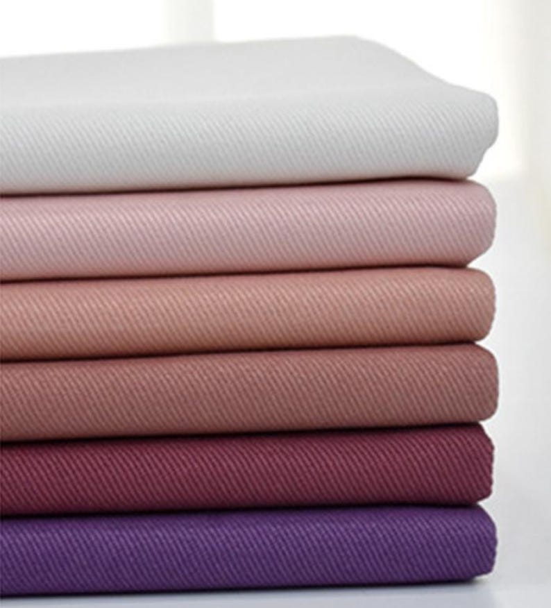 May include: A stack of six folded fabric pieces arranged in a gradient from white to light pink, dusty rose, mauve, deep berry, and dark purple. Each piece of fabric features a visible diagonal twill weave texture throughout the entire surface.