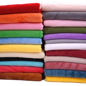 May include: A stack of folded fabric squares in a wide variety of solid colours including red, orange, pink, green, blue, yellow, cream, black, white, grey, purple, maroon, and brown. Each piece of fabric has a soft, fuzzy, plush texture.