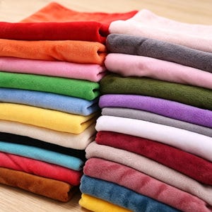 May include: A stack of folded fabric squares in a variety of solid colors including red, orange, pink, green, blue, yellow, black, white, brown, gray, and purple. Each piece of fabric features a soft, fuzzy, plush texture throughout the pile.