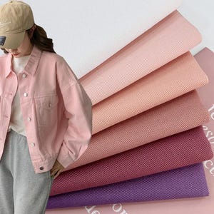 May include: A light pink denim jacket with a collar, long sleeves, and silver-toned metal buttons. Beside the jacket are stacked layers of fabric in various shades including white, light pink, peach, dusty rose, dark pink, and purple.