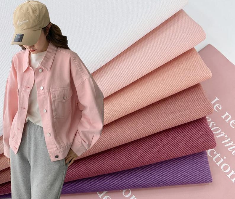 May include: A light pink denim jacket with a collar, long sleeves, and silver-toned metal buttons. Beside the jacket are stacked layers of fabric in various shades including white, light pink, peach, dusty rose, dark pink, and purple.