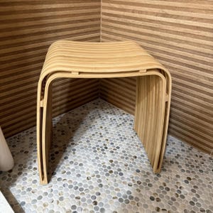 May include: A light brown bamboo stool featuring a curved, slatted seat and matching side panels. The stool is constructed from multiple thin, horizontal strips of wood that form a continuous, rounded shape with open sides and a smooth finish.