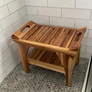 May include: A rectangular wooden shower bench featuring a slatted top surface with integrated handles on each side and a lower storage shelf. The bench is constructed from natural brown wood with visible grain patterns and four sturdy legs.