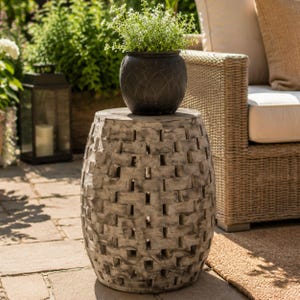 May include: A barrel-shaped garden stool with a weathered gray finish and a repeating cutout pattern. The stool has a flat circular top surface and a textured exterior. A dark charcoal round planter with a small green plant sits on top.