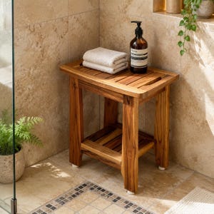 May include: A rectangular wooden shower bench with a slatted top and a lower shelf. The bench features four sturdy legs with white protective feet. A folded white towel and a brown amber-colored bottle with a black pump dispenser sit on top.