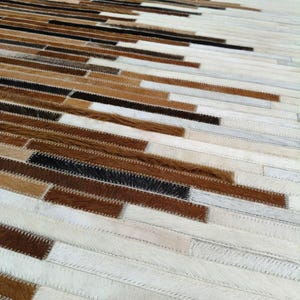 May include: A rectangular area rug constructed from numerous narrow, horizontal strips of cowhide leather. The strips feature a color palette of white, cream, tan, brown, and black, arranged in a staggered, layered pattern with visible stitching.