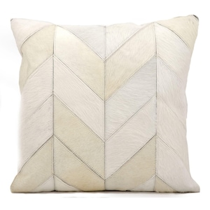 May include: A square throw pillow featuring a chevron pattern constructed from stitched panels of off-white and cream-colored hair-on hide. The textured surface displays a repeating geometric arrangement of angled strips.
