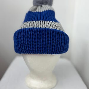 Puede incluir: Un gorro de punto azul, gris y blanco con un pompón gris en la parte superior. El sombrero se exhibe en una cabeza de maniquí blanca. El sombrero tiene un borde grueso y acanalado en azul, con rayas grises y azules alternas.