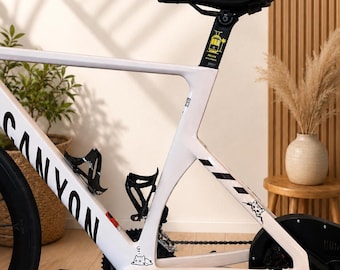 Könnte beinhalten: Ein weißer Canyon Rennradrahmen auf einem schwarzen Tacx Rollentrainer. Der Rahmen hat schwarze Schriftzüge, eine kleine Katzen-Grafik und einen gelben Aufkleber an der Sattelstütze, der eine Straßenbahn mit dem Text Soller Mallorca zeigt.