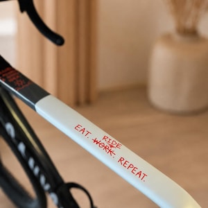 Könnte beinhalten: Ein weißer Fahrradrahmen mit rotem Text, der EAT. RIDE. WORK. REPEAT. lautet. Der Rahmen umfasst einen schwarzen Lenker und ein teilweise sichtbares schwarzes Rad im unteren Bereich. Das Design ist schlicht mit einer glatten Oberfläche am Oberrohr.