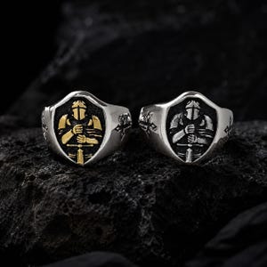 Könnte beinhalten: Zwei silberfarbene Ringe mit schildförmiger Vorderseite, die einen gepanzerten Ritter mit Schwert zeigen. Ein Ring hat eine goldfarbene Ritterfigur vor schwarzem Hintergrund, während der andere einen silberfarbenen Ritter auf schwarzem Grund zeigt.