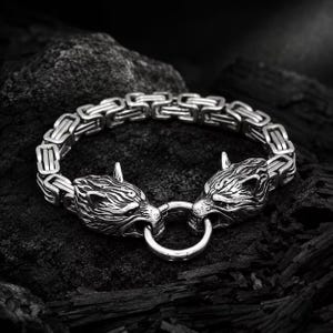 Può includere: Un bracciale in metallo color argento caratterizzato da una spessa catena a maglia bizantina intrecciata. Le due estremità del bracciale terminano con sculture dettagliate di teste di lupo contrapposte, unite da una chiusura ad anello metallico.