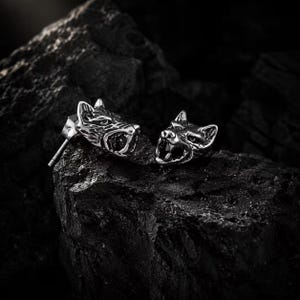 May include: A pair of silver-toned stud earrings shaped like snarling wolf heads. Each earring features detailed facial features, including pointed ears, bared teeth, and a textured metallic finish, designed as a post-back style accessory.