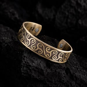 Könnte beinhalten: Ein messingfarbenes Metall-Armband mit einem sich wiederholenden, geprägten Triskelion-Spiralmuster. Die Innenseite des offenen Armreifs hat eine gebürstete Oberfläche und enthält zwei kleine, kreisförmige Magnete an den Enden.