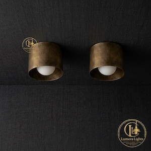 May include: Two cylindrical flush mount ceiling lights featuring a brushed antique brass finish. Each fixture houses a single spherical white glass globe bulb centered within the metal housing. The Lumera Lights logo is visible in the corners.