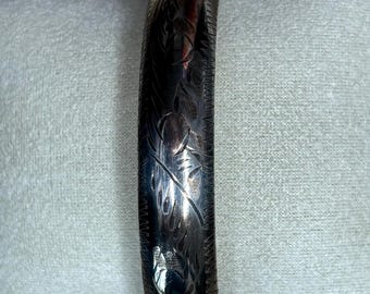May include: A silver-toned metal bangle bracelet featuring an intricate, engraved floral pattern across its curved surface. The bracelet has a polished finish with dark, recessed lines highlighting the detailed botanical etchings and a hinged clasp.