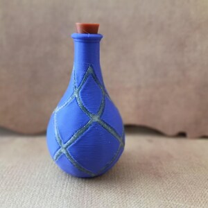 Pode incluir: Uma pequena garrafa azul com corpo arredondado e gargalo estreito, apresentando um padrão de grade texturizado em forma de losango na superfície. A garrafa é rematada com uma rolha cilíndrica castanha simples que encaixa na abertura.