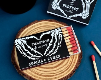 Può includere: Una scatola di fiammiferi rettangolare con una copertina nera raffigurante mani di scheletro bianche che formano un cuore, il testo "TILL DEATH do us part" e "SOPHIA & ETHAN". La scatola è socchiusa e rivela fiammiferi con punte rosse.