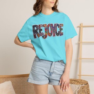 Peut inclure: Un t-shirt turquoise à manches courtes avec le mot REJOYCE imprimé sur la poitrine. Les lettres sont remplies d'un motif patchwork coloré et complexe composé de diverses formes géométriques, de motifs floraux et de petites icônes dans des tons de rouge, jaune, bleu et vert.