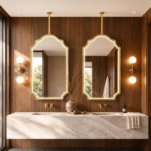 May include: Two wall-mounted mirrors with gold frames and integrated LED lighting, suspended by vertical gold rods. The mirrors are positioned above a long, floating stone vanity with two integrated sinks, gold faucets, and wall-mounted globe lights.