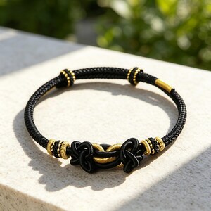 May include: A black cord bracelet featuring intricate woven knots in the center. The design includes metallic gold-toned accents and small cylindrical beads integrated along the band, creating a structured and detailed circular wrist accessory.
