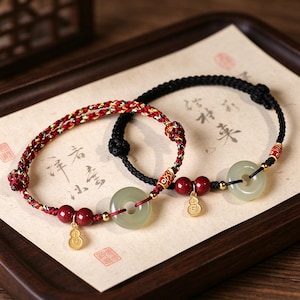 May include: Two bracelets featuring braided cords in red, yellow, and black. Each bracelet includes a pale green jade-like ring, two deep red beads, a small gold-toned gourd charm, and a decorative gold-toned bead with a red fish pattern.