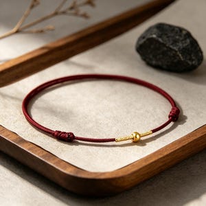 Puede incluir: Una pulsera fina de cordón rojo oscuro con una cuenta esférica dorada pulida en el centro, flanqueada por detalles cilíndricos dorados texturizados. La pulsera incluye extremos de cordón anudados ajustables para un ajuste seguro.