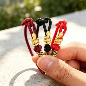 Könnte beinhalten: Drei Kordelarmbänder mit einem zentralen goldfarbenen Schlangenanhänger, der sich um das Seil windet. Die Armbänder bestehen aus roter und schwarzer geflochtener Kordel, jeweils mit einem Knoten über der Metallschlange und einem gewickelten Ende.