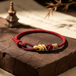 May include: A red cord bracelet featuring a gold-colored snake charm. The bracelet is secured with a knot and includes a decorative section wrapped in thin, multi-colored threads near the clasp, creating a textured and detailed accessory.