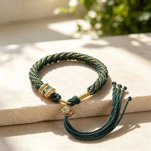 May include: A dark green and gold twisted cord bracelet featuring a cylindrical gold-tone bead with green enamel and engraved text, a spherical gold-tone bead with red and green accents, and a long tassel made of multiple dark green cords.